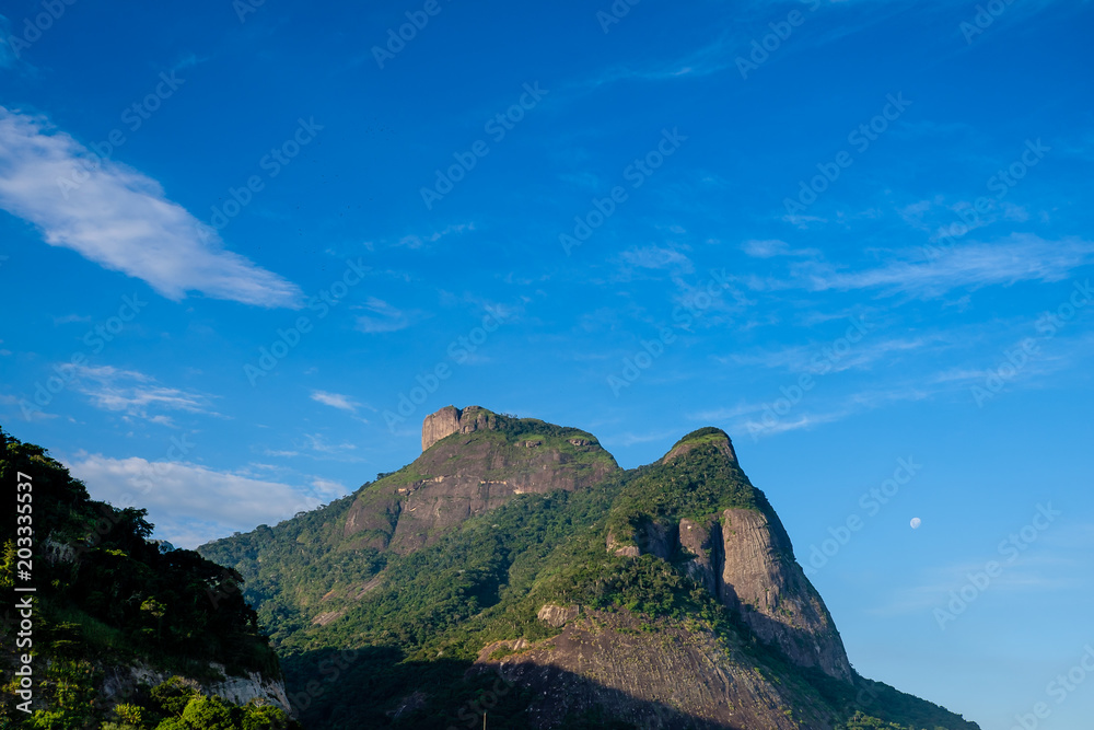 Fototapeta premium View of the Gavea Stone, seen from the street with houses on the hill during late afternoon. Barra da Tijuca, Rio de Janeiro