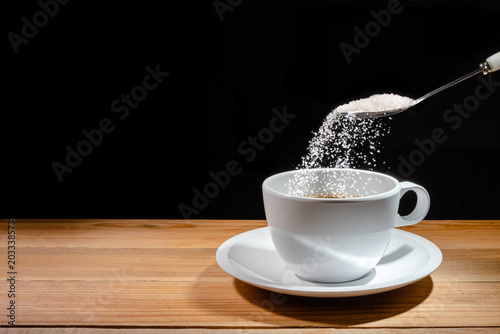 spoon is pouring sugar on coffee cup with saucer on wooden table on black background