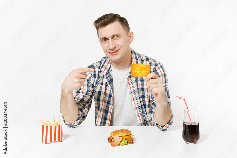 Handsome young man sitting at table with credit card, burger, french fries, cola in glass isolated on white background. Proper nutrition or American classic fast food. Advertising area with copy space Handsome young man sitting at table with credit card, burger, french fries, cola in glass isolated on white background. Proper nutrition or American classic fast food. Advertising area with copy space