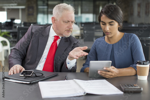 Frowning boss annoyed with managers report. Young Indian woman in casual wear showing financial data on tablet to mid adult man in modern business space. Teamwork and cooperation concept