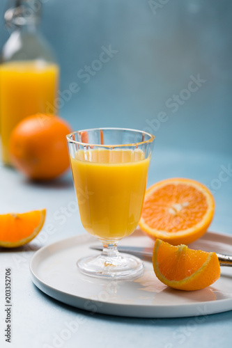 Glass of fresh orange juice with orange slices on white plate, blue background, cold summer refreshing nutritional beverage, blue orange color contrast, vitamin C healthy drink, copy space