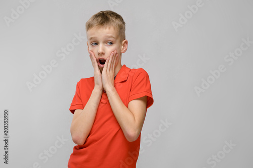 Portrait of little blonde surprised astonished caucasian boy in orange t-shirt with open mouth on gray background