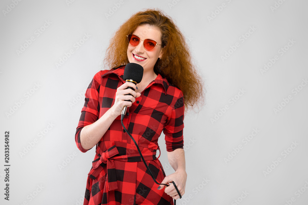 Portrait of beautiful redhead happy young woman in sunglasses smiling holding microphone on gray background Portrait of beautiful redhead happy young woman in sunglasses smiling holding microphone on gray background