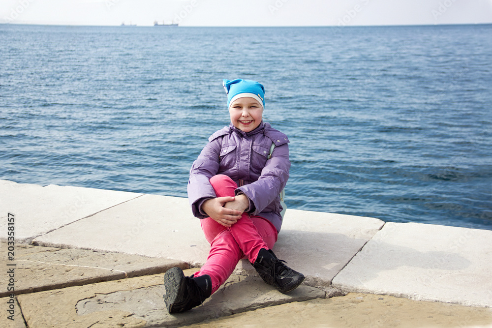 young little happy sunny girl in blue hat and pink pants is sitting fashion and posing with  hands on leg on the blue sea background and smiling