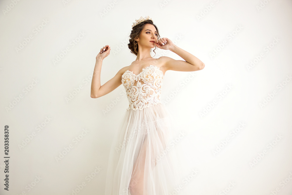 young brunette sexy girl in a white wedding boudoir dress and gold crown in hairstyle is standing with hands near head like swan princess on a white wall background young brunette sexy girl in a white wedding boudoir dress and gold crown in hairstyle is standing with hands near head like swan princess on a white wall background