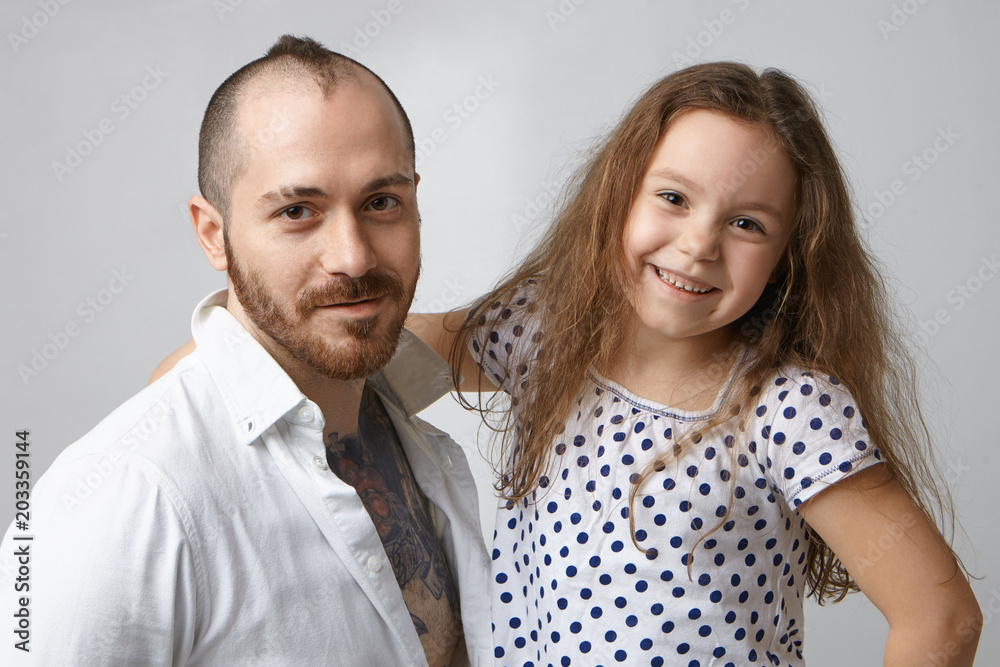 Portrait of happy stylish modern young father with beard and creative hairstyle posing in studio with his cheerful charming daughter who is embracing him and looking at camera with joyful smile Portrait of happy stylish modern young father with beard and creative hairstyle posing in studio with his cheerful charming daughter who is embracing him and looking at camera with joyful smile