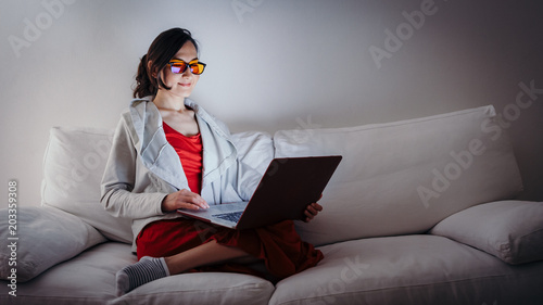 Young woman with blue light blocking glasses (yellow lenses) working with laptop on white sofa