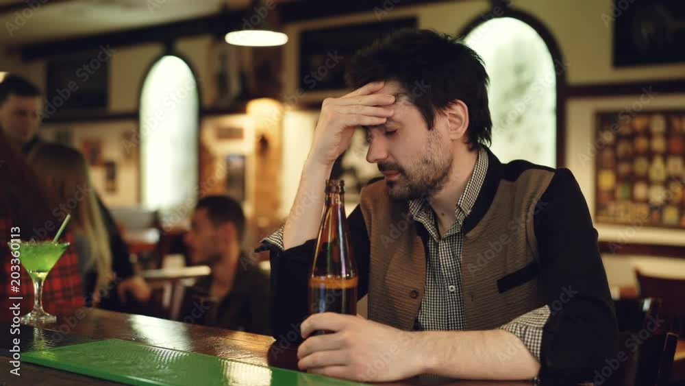 Upset unshaven guy is sitting at bar counter with bottle of beer, sighing and touching his face in despair. He is watching young people having fun and frowning. Upset unshaven guy is sitting at bar counter with bottle of beer, sighing and touching his face in despair. He is watching young people having fun and frowning.