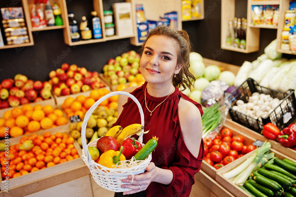 Girl in red holding different fruit and vegetables at basket on fruits store.