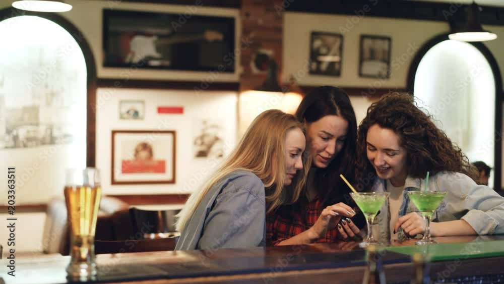 Girls are using smartphone, laughing and talking while sitting in bar together. Girls in casual clothing are watching screen and chatting. Modern technologies for fun concept. Girls are using smartphone, laughing and talking while sitting in bar together. Girls in casual clothing are watching screen and chatting. Modern technologies for fun concept.