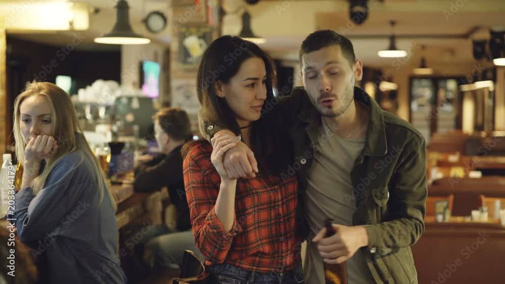 Dolly shot of young woman helping her drunk boyfriend leave bar. Handsome bearded man is holding beer bottle and telling something to his girlfriend.