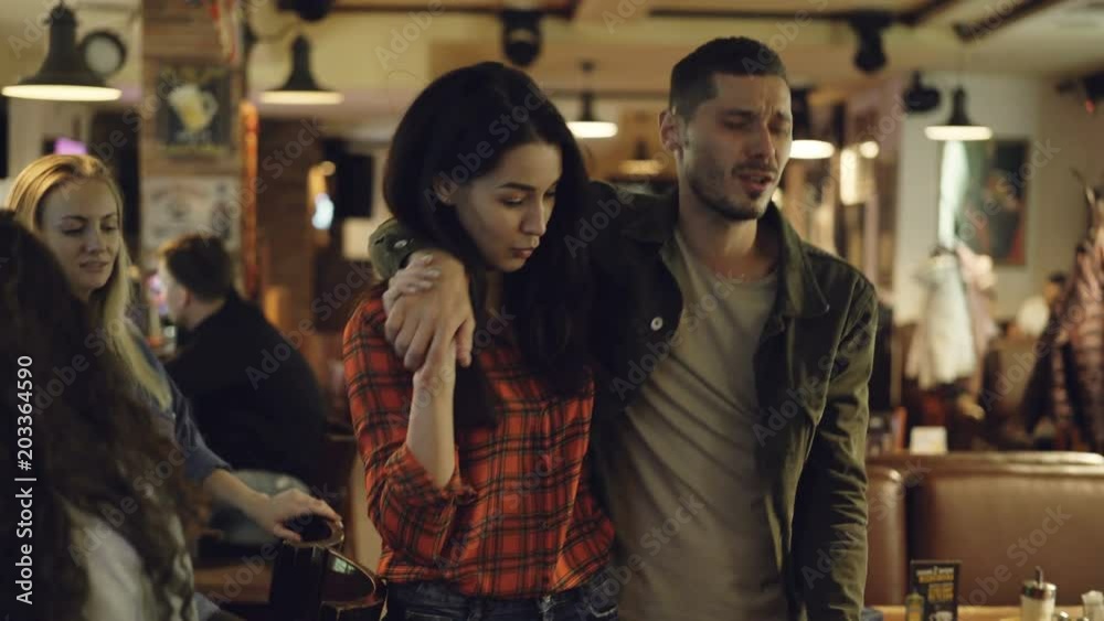 Young brunette is leading her drunk husband out of bar. Unshaven handsome young man is talking to his upset wife trying to explain himself. Alcoholism ruining family concept.