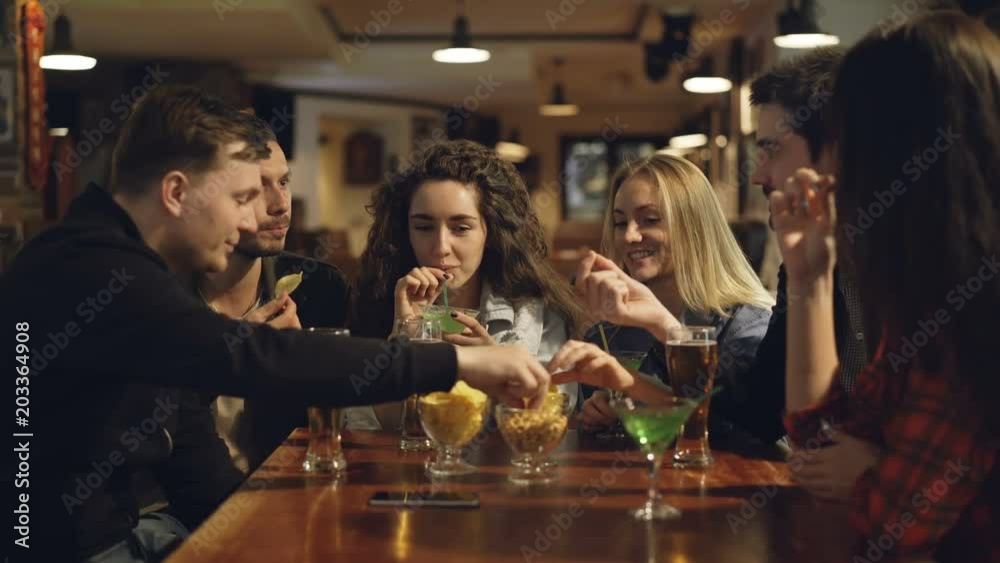 Group of friends are drinking, eating snacks and talking while sitting at table in bar. Young people enjoying themselves and communicating in pub concept.
