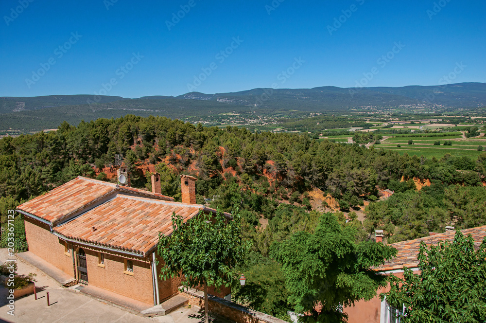 Panoramic view of the fields and hills of Provence from the city center of Roussillon, with stone houses, roofs and trees in the foreground. Vaucluse department, Provence region, southeastern France. 