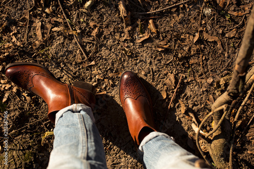 Brown shiny leather womens chelsea boots on woman legs with blue jeans in autumn forest or park.