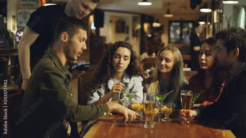 Handsome bearded guy is paying with smartphone in pub while having fun with friends. Friendly waiter, table with drinks and snacks, nice bar interior are visible.