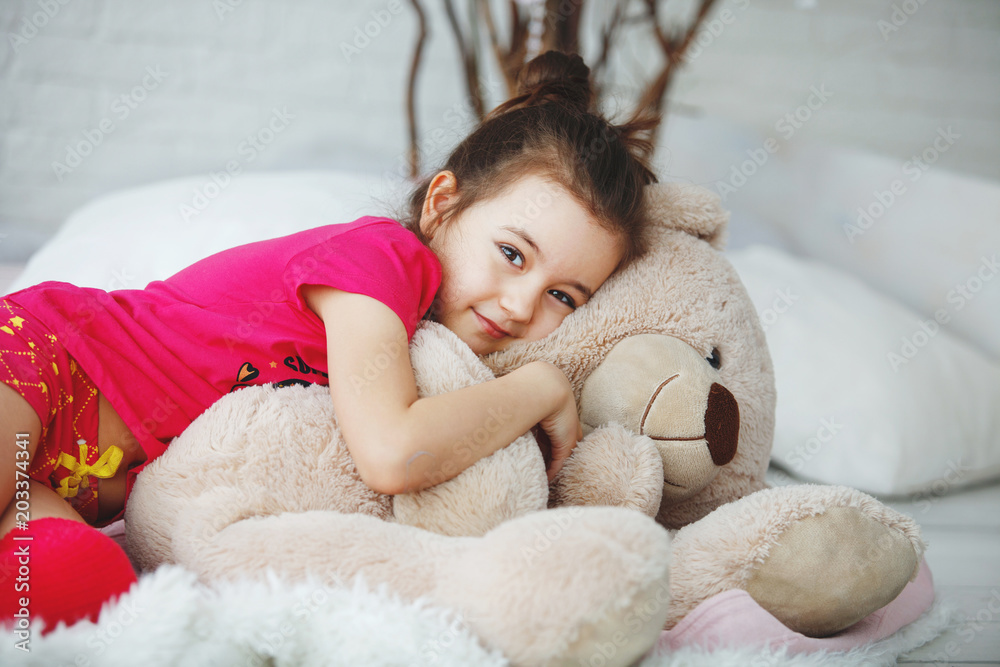 A young long-haired girl in a pink T-shirt and shorts and golfs lies, hugs with a toy bear in bed with cushions and tree branches A young long-haired girl in a pink T-shirt and shorts and golfs lies, hugs with a toy bear in bed with cushions and tree branches