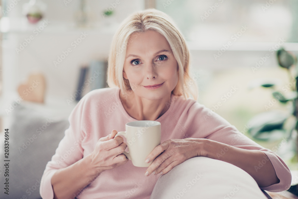 Modern, stylish, caucasian, aged, woman having cup of coffee in the morning, sitting on sofa in living room at home, looking at camera Modern, stylish, caucasian, aged, woman having cup of coffee in the morning, sitting on sofa in living room at home, looking at camera