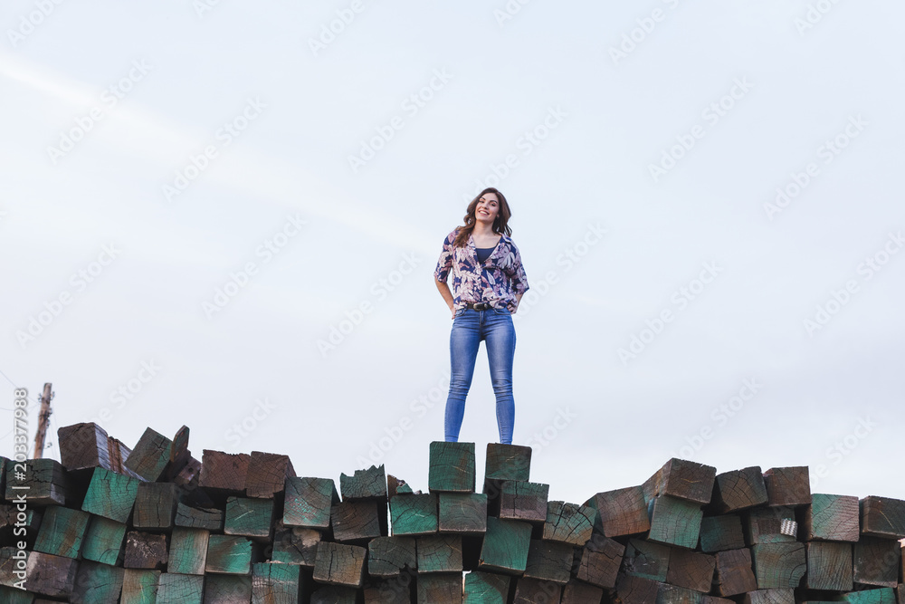 Obraz premium portrait of a young beautiful woman wearing casual clothes, standing on a mountain of green wood blocks background and smiling. Outdoors lifestyle.