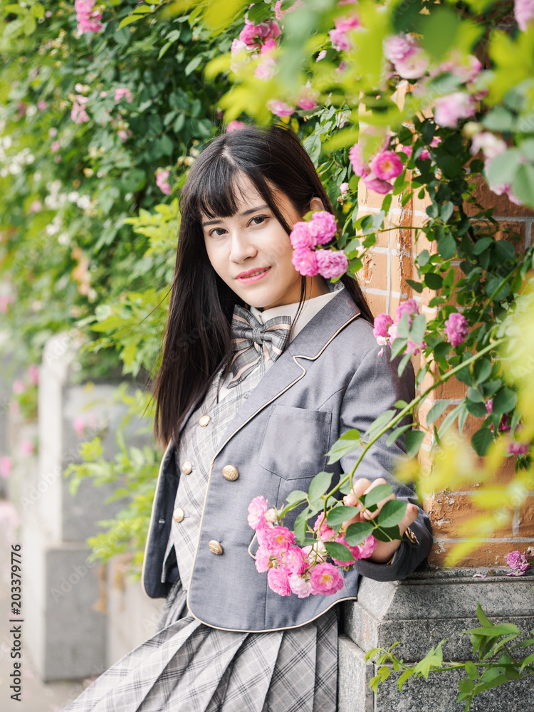 Fototapeta premium Outdoor portrait of beautiful young Chinese girl in school uniform japanese style smiling among pink rose flowers wall in spring garden, Perfect Skin. Professional Make-up.