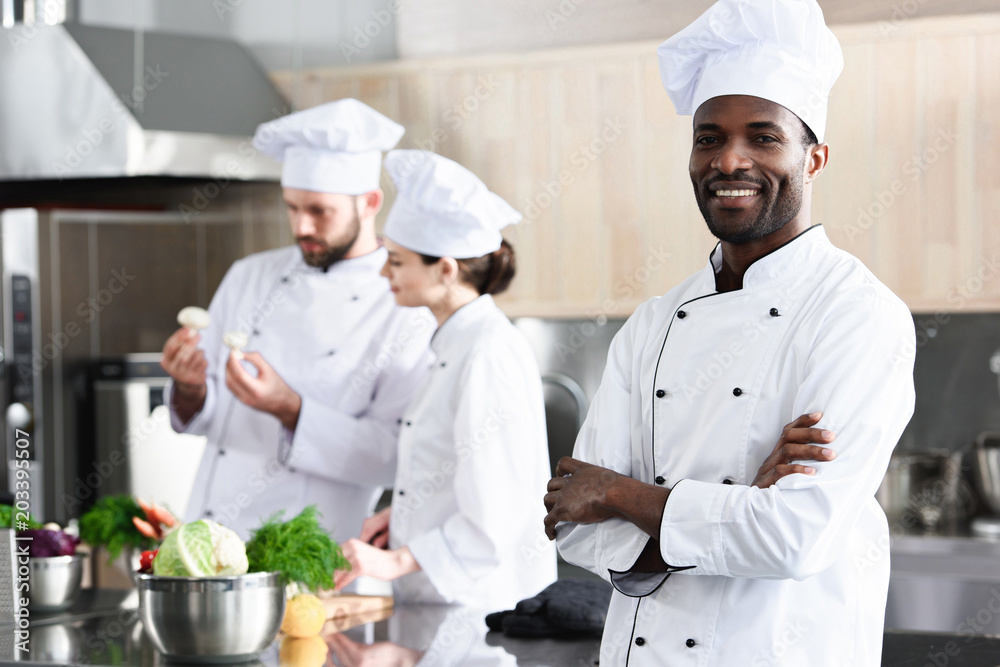 African american chef standing with folded arms in front of his colleagues on kitchen