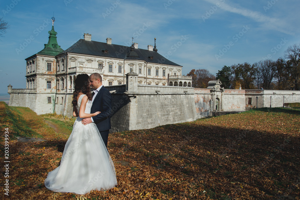 Wedding couple is kissing outdoors in sunny autumn day. Beautiful nature and renaissance palace on background. Bride is satin lace dress is holding groom hand. Old castle architecture.