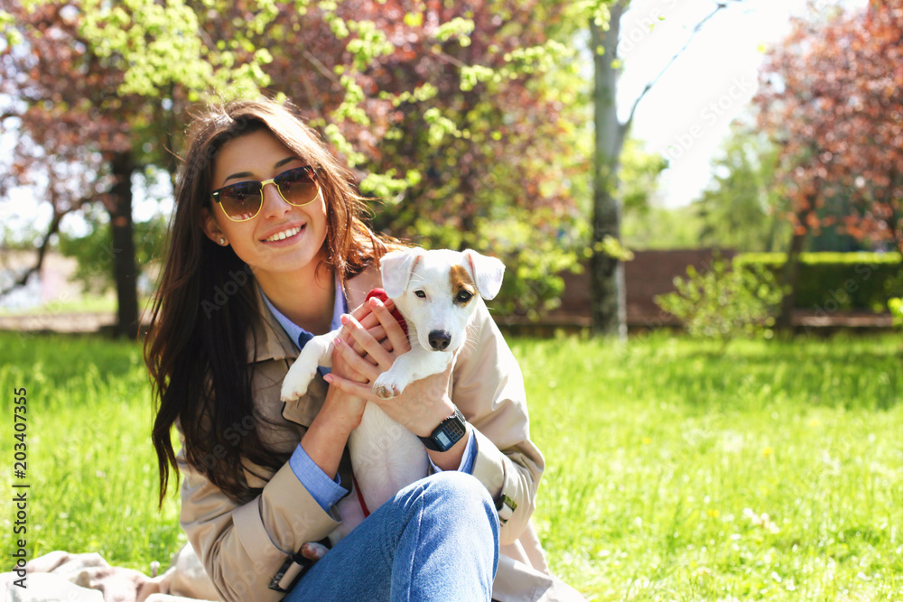 Portrait of attractive young woman hugging cute jack russell terrier puppy in park, green lawn, foliage background. Hipster female in sunglasses smiling, pets her dog on grass. Close up, copy space.