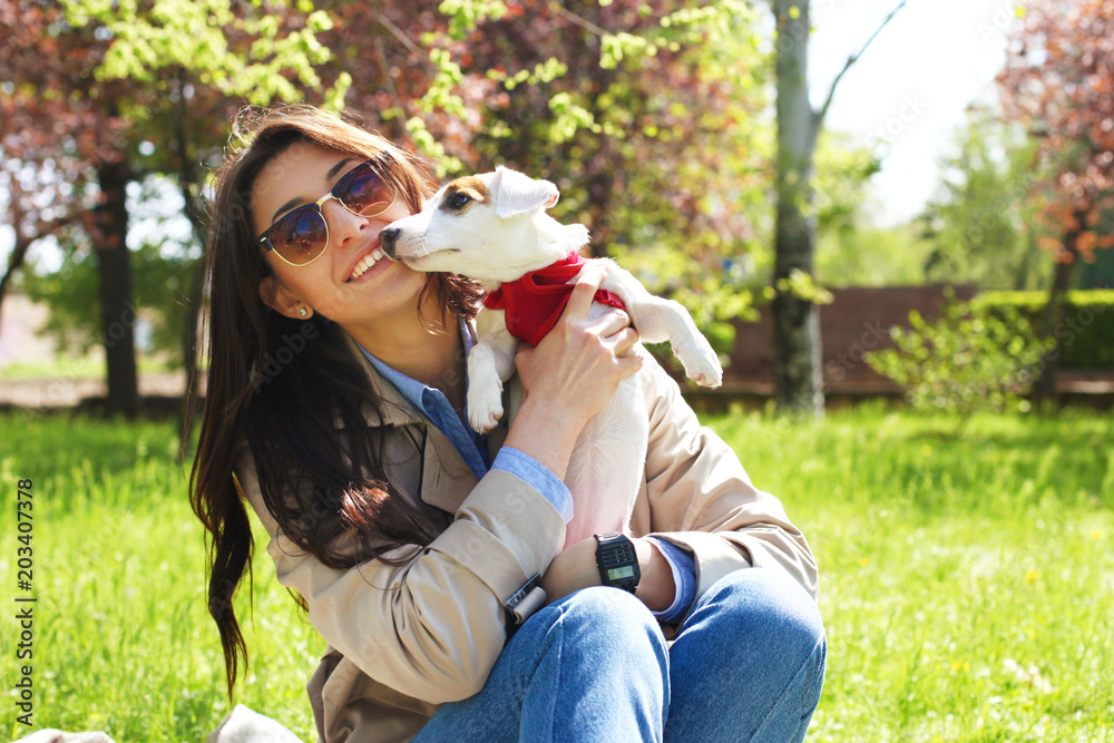 Naklejka premium Portrait of attractive young woman hugging cute jack russell terrier puppy in park, green lawn, foliage background. Hipster female in sunglasses smiling, pets her dog on grass. Close up, copy space.