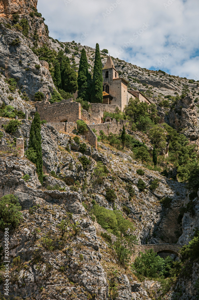 View of the church Notre-Dame de Beauvoir amid the cliffs and houses of the charming village Moustiers-Sainte-Marie. In the Alpes-de-Haute-Provence department, Provence region, southeastern France