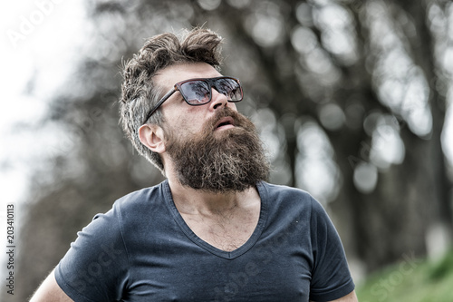 Barbershop and style concept. Man with beard and mustache on strict face, nature background, defocused. Hipster with beard looks confident while standing outdoors. Bearded man wears modern sunglasses.