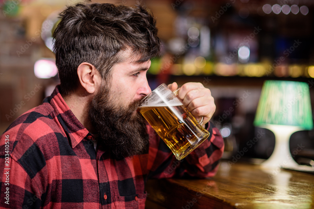 Guy spend leisure in bar, defocused background. Hipster with beard holds glass with beer, drinks, enjoy fresh taste. Relaxation concept. Man with tired or calm face sit in bar or pub near bar counter. Guy spend leisure in bar, defocused background. Hipster with beard holds glass with beer, drinks, enjoy fresh taste. Relaxation concept. Man with tired or calm face sit in bar or pub near bar counter.