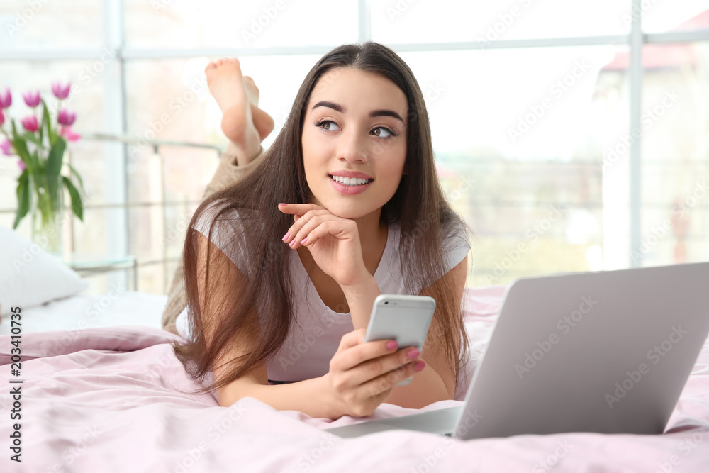 Attractive young woman using mobile phone and laptop on bed at home Attractive young woman using mobile phone and laptop on bed at home