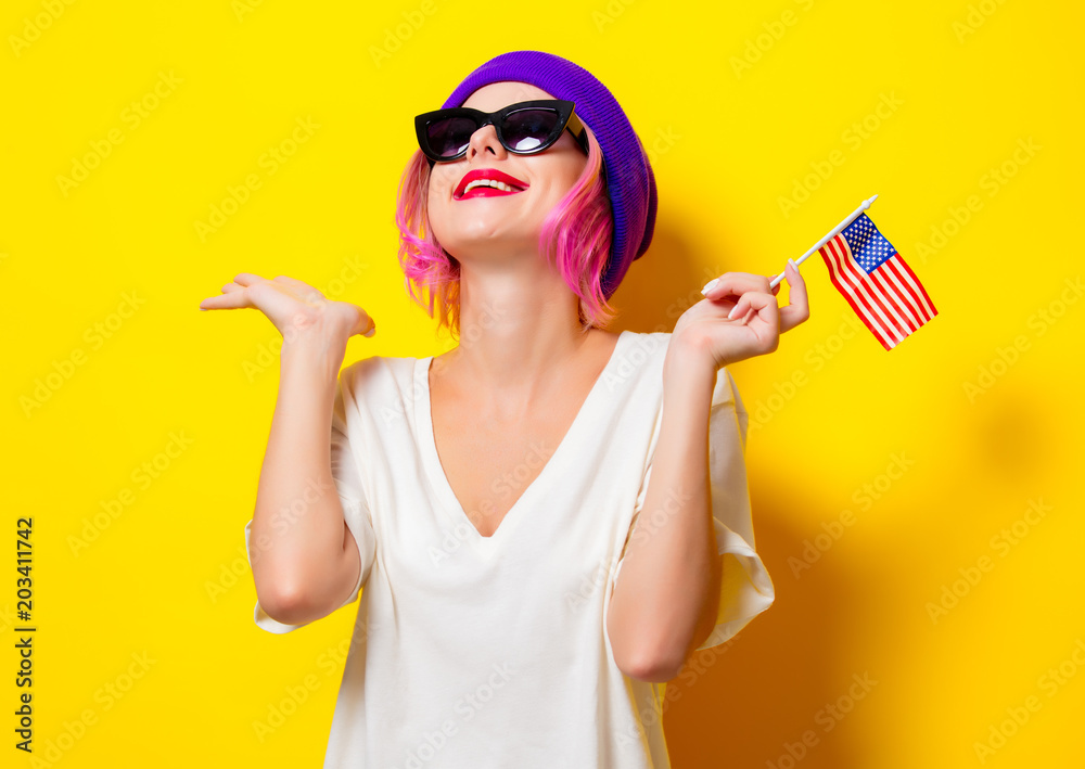 Young girl with pink hair in purple hat and sunglasses holding United States flag on yellow background