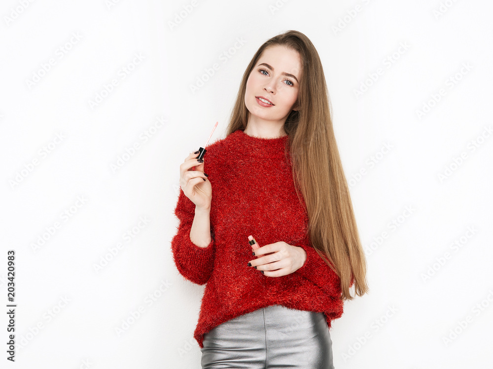 Spectacular blonde woman in red blouse silver leather pants posing with lip glow balm in front of white wall. Graceful girl gorgeous long hair having fun on photoshoot.