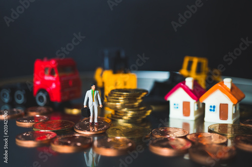 Miniature people  businessman standing with golden stack coins on construction vehicle background using as business and construction concept