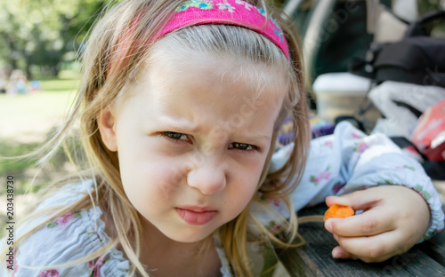 Grumpy Angry Girl Child Holding Carrot Vegetable at Picnic Table - Refusing to Eat 5-a-Day or Healthy-Eating