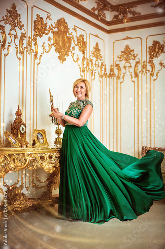 Beautiful blonde woman in long green dress is standing by wall in luxurious room
