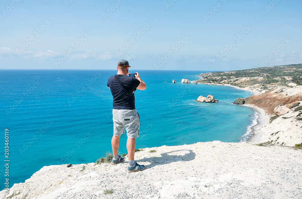 Travel man taking photo with mobile phone standing near sea. Hiking man standing on top of mountain and making photo. Travel, vacation concept. Cyprus, Mediterranean Sea.