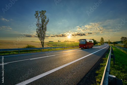 Red bus traveling on asphalt road around farm fields in rural landscape at sunset © am