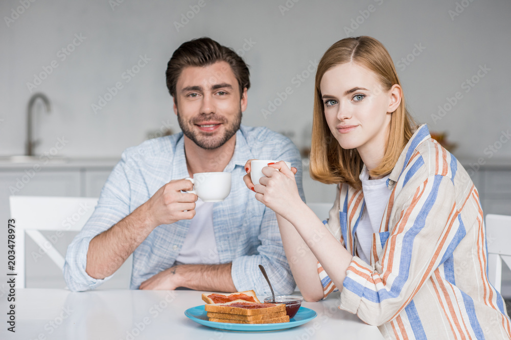 smiling young couple with coffee cups having breakfast at table in kitchen smiling young couple with coffee cups having breakfast at table in kitchen
