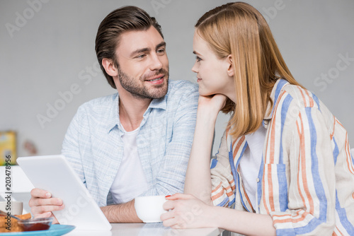 attractive young woman with coffee cup and boyfriend with digital tablet looking at each other