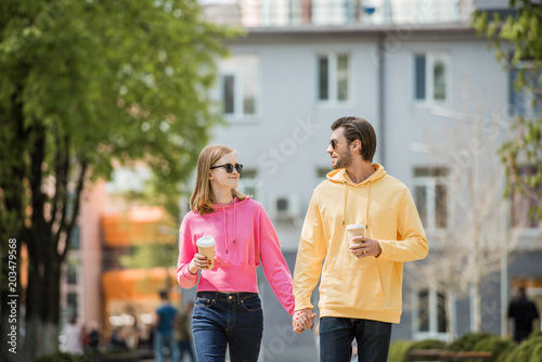 Stylish young couple in sunglasses with coffee cups holding hands and walking on street
