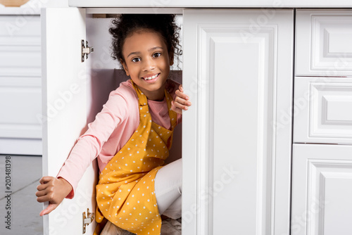 smiling african american child in apron hiding on kitchen