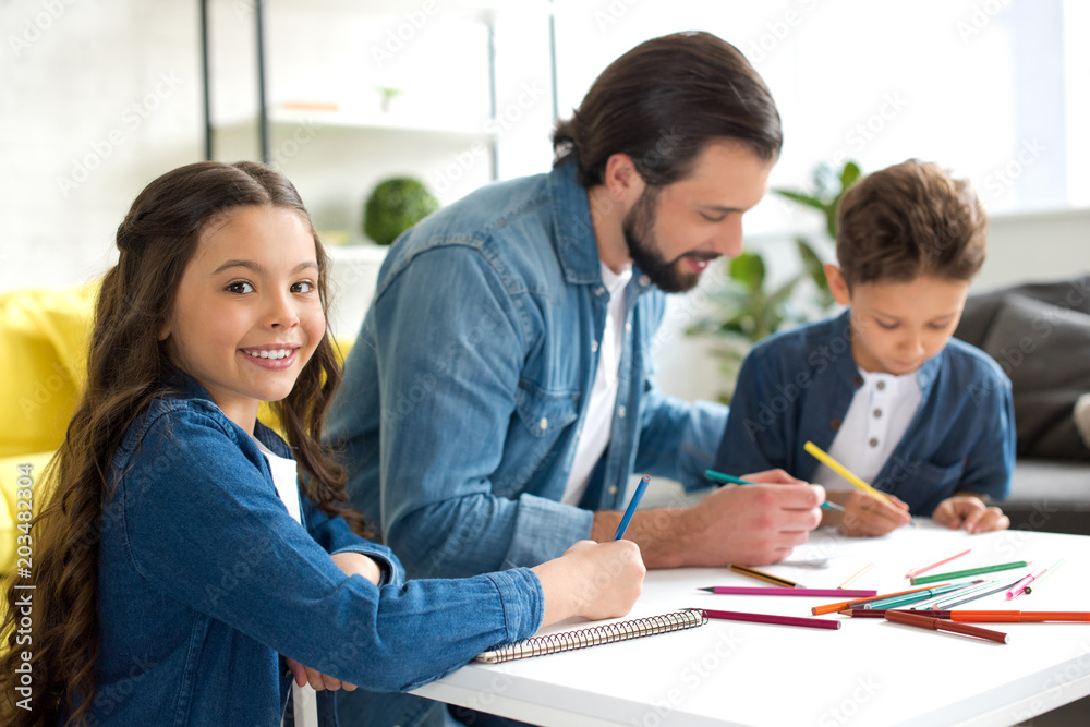 adorable child smiling at camera while drawing with father and brother at home adorable child smiling at camera while drawing with father and brother at home