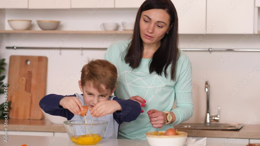 Mother and son are standing by the kitchen counter; mother teaches son to make an omelette; son cracks eggs by himself; Mom praises kid