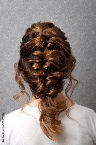 Hairstyle Greek braid on the head of a brown-haired woman back view close-up on a gray background.