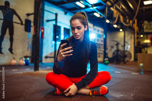 Attractive young brunette is sitting on floor in a gym and using her smartphone