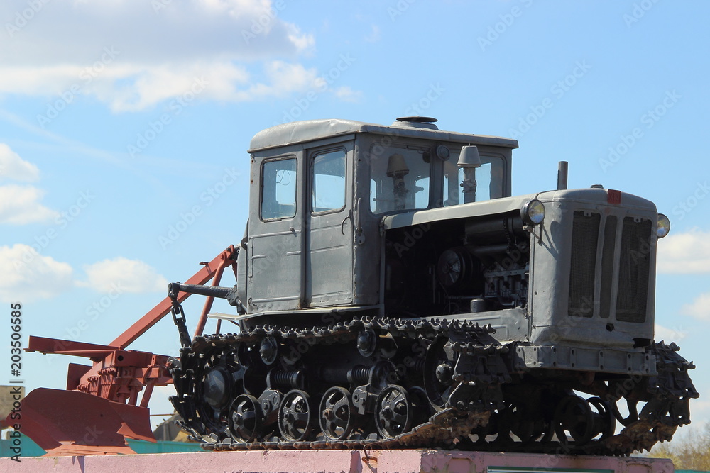 Grey Soviet crawler tractor with plow on pedestal in spring on blue sky background with clouds Grey Soviet crawler tractor with plow on pedestal in spring on blue sky background with clouds