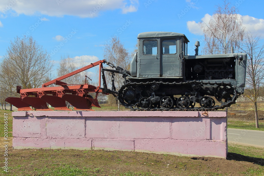 Fototapeta premium Grey Soviet crawler tractor with plow on pedestal in spring on blue sky background with clouds
