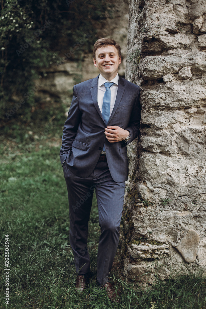 Joyful groom in elegant blue suit smiling and posing near old castle wall, handsome businessman posing outdoors, newlywed groom portrait concept Joyful groom in elegant blue suit smiling and posing near old castle wall, handsome businessman posing outdoors, newlywed groom portrait concept